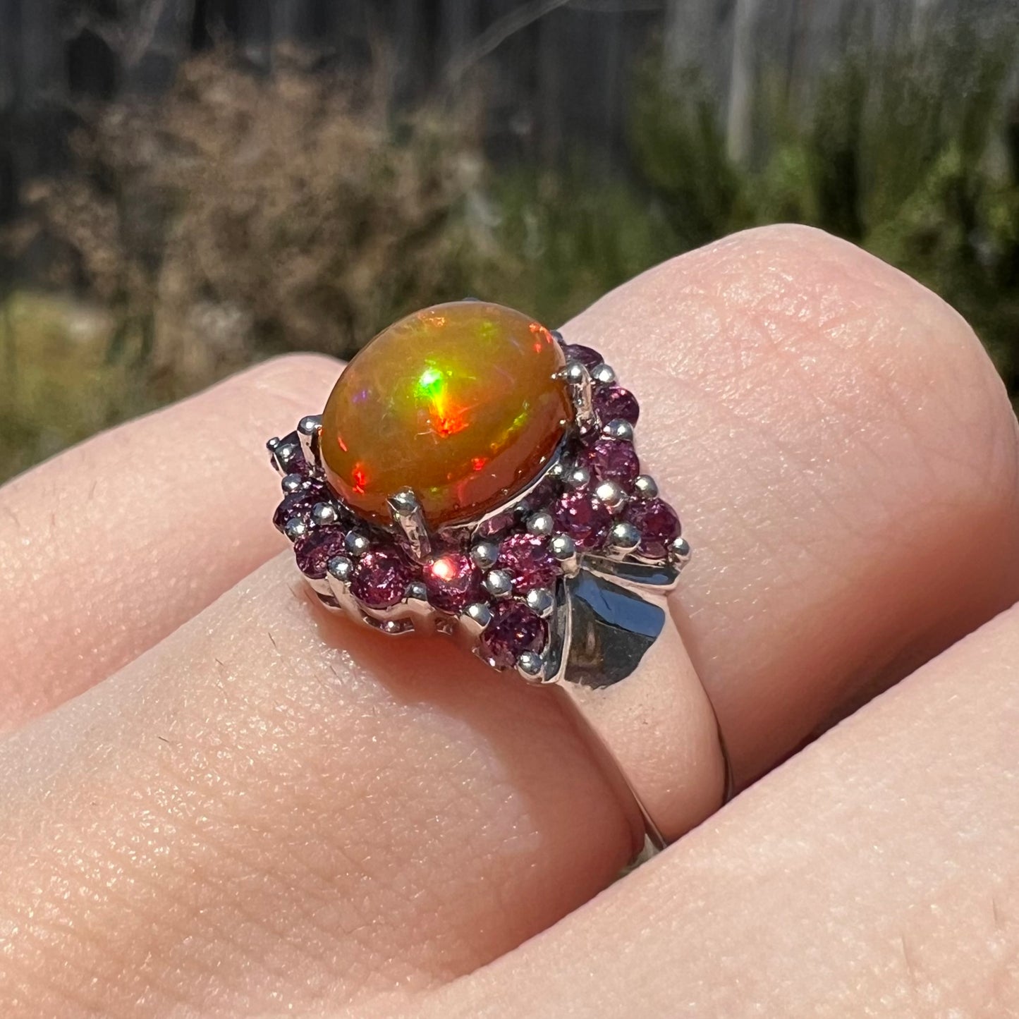 Angled close-up of fire opal and purple garnet ring worn on finger in sunlight, the opal has iridescent red and green colors