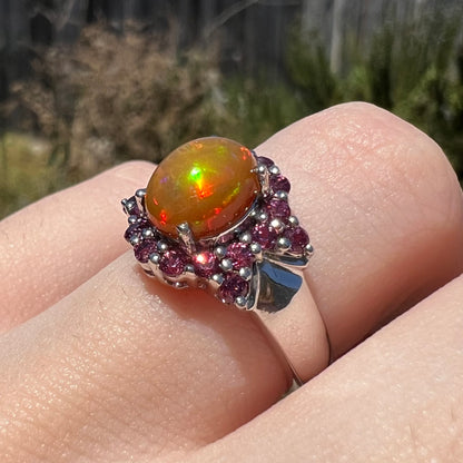 Angled close-up of fire opal and purple garnet ring worn on finger in sunlight, the opal has iridescent red and green colors