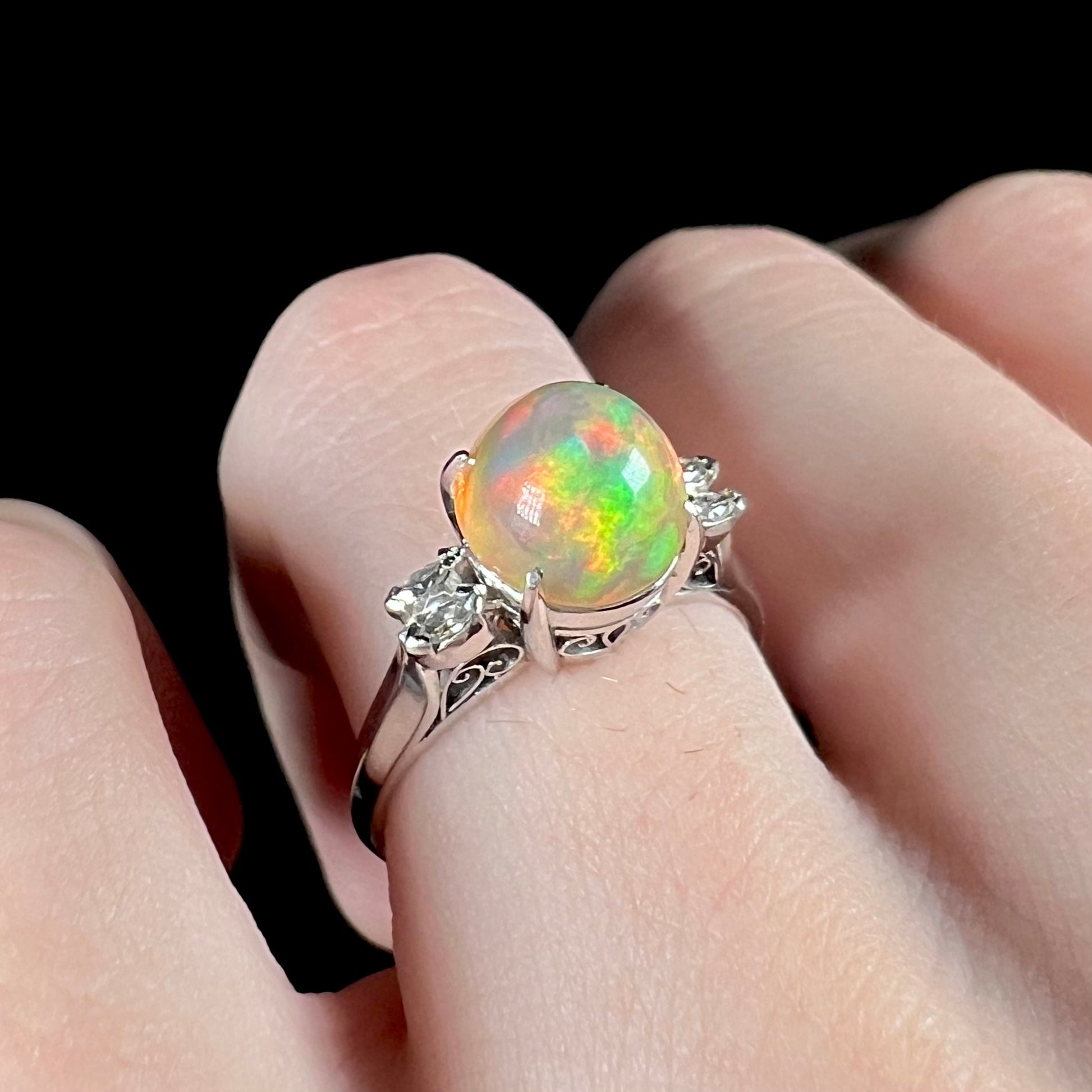 Angled close-up of a fire opal engagement ring worn on a finger, featuring a 2.28ct Mexican fire opal with red and green play-of-color and platinum filigree.