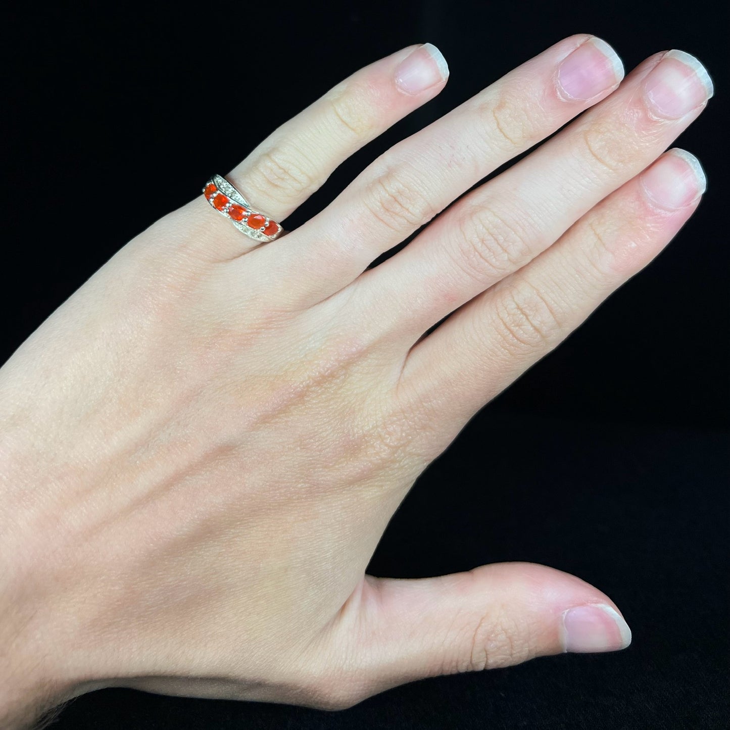 Hand wearing a silver fire opal ring on its pinky finger against a black background.