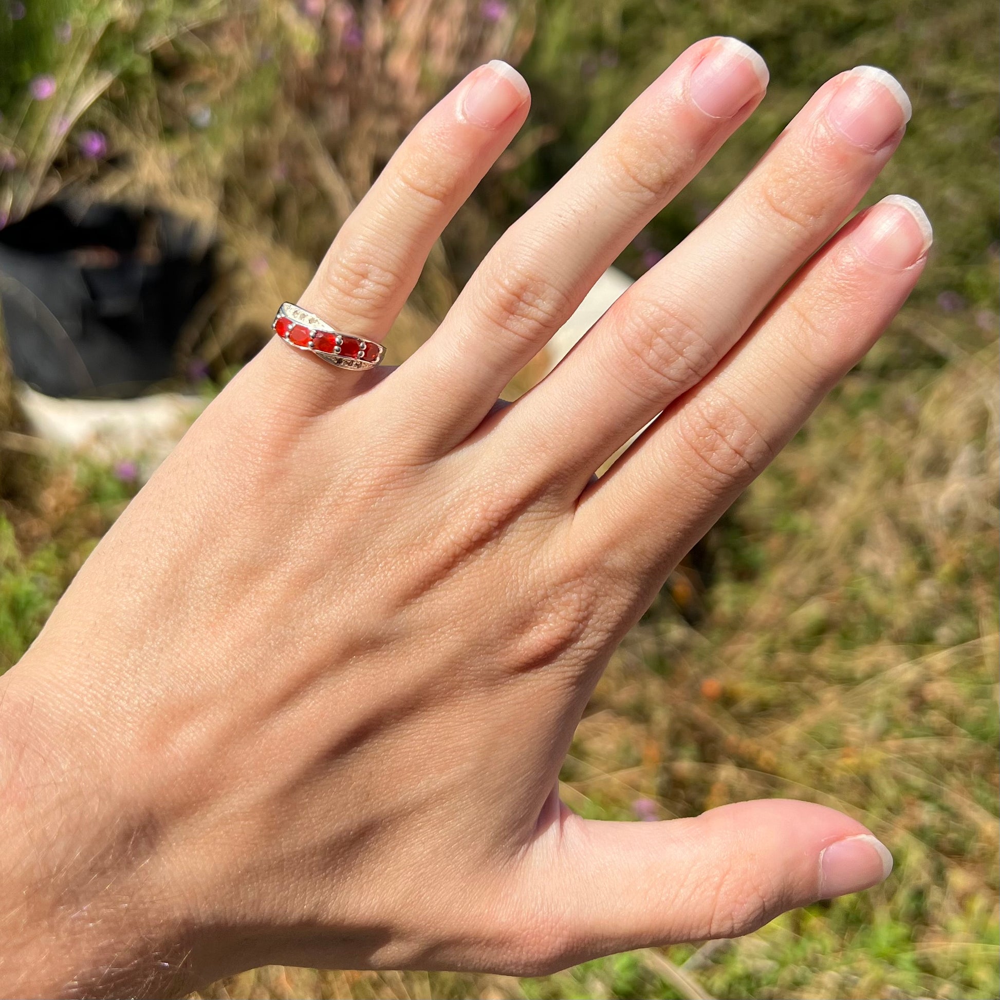 Hand wearing silver fire opal and diamond ring on pinky finger in natural sunlight against blurred outdoor background.