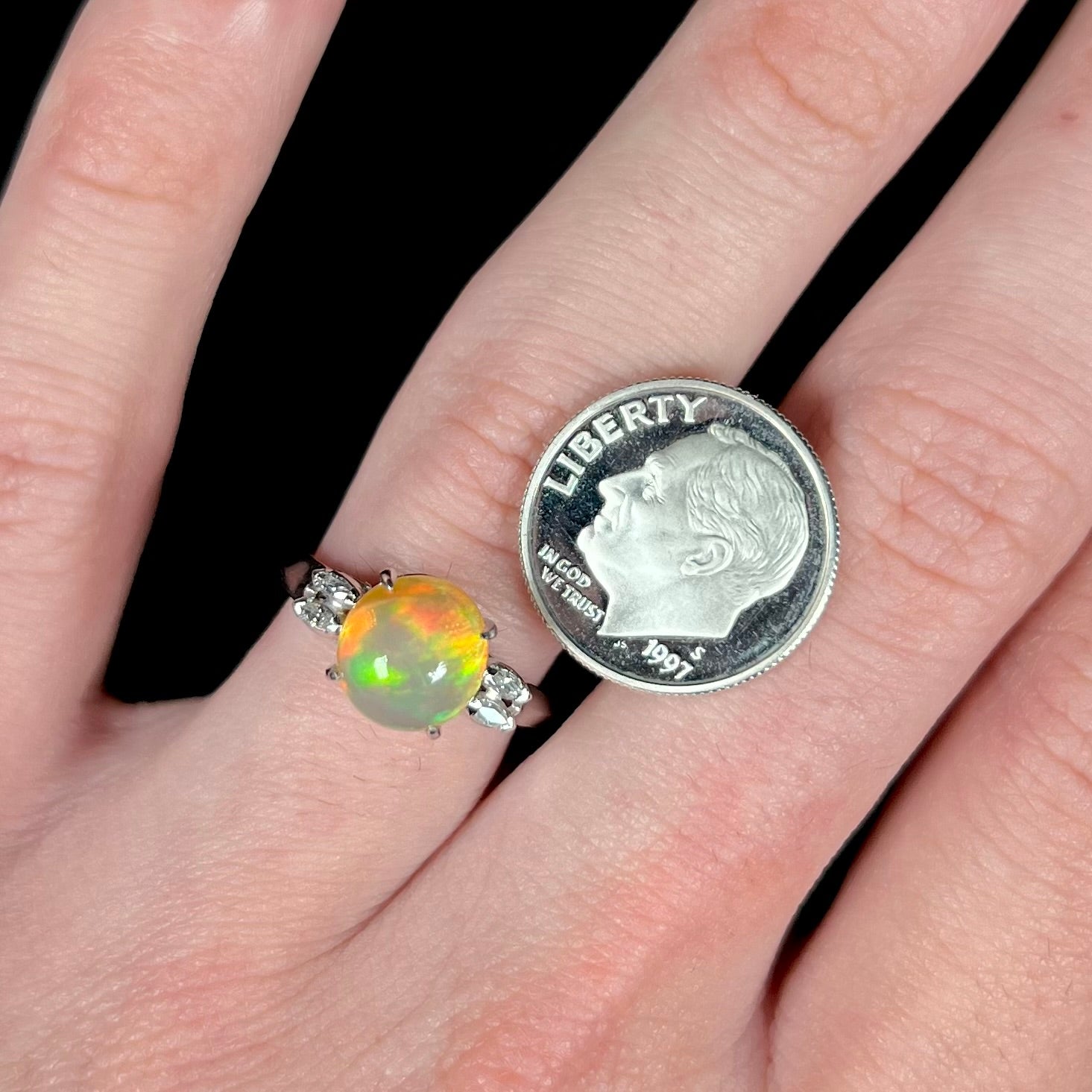 A platinum fire opal and diamond ring featuring a round 2.28ct Mexican fire opal, on a hand next to a US dime coin.