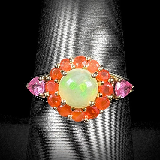 A yellow gold ring mounted with a round Ethiopian opal in a halo of faceted Mexican fire opals.  Two pear shaped pink tourmalines accent the sides of the ring.