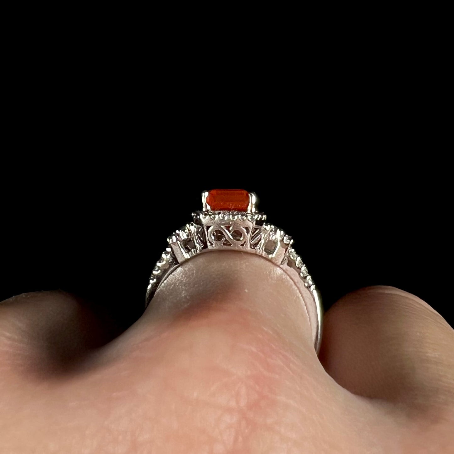 Close-up of the side gallery view of a white gold fire opal ring with a cast filigree design, worn on a finger against a black background.