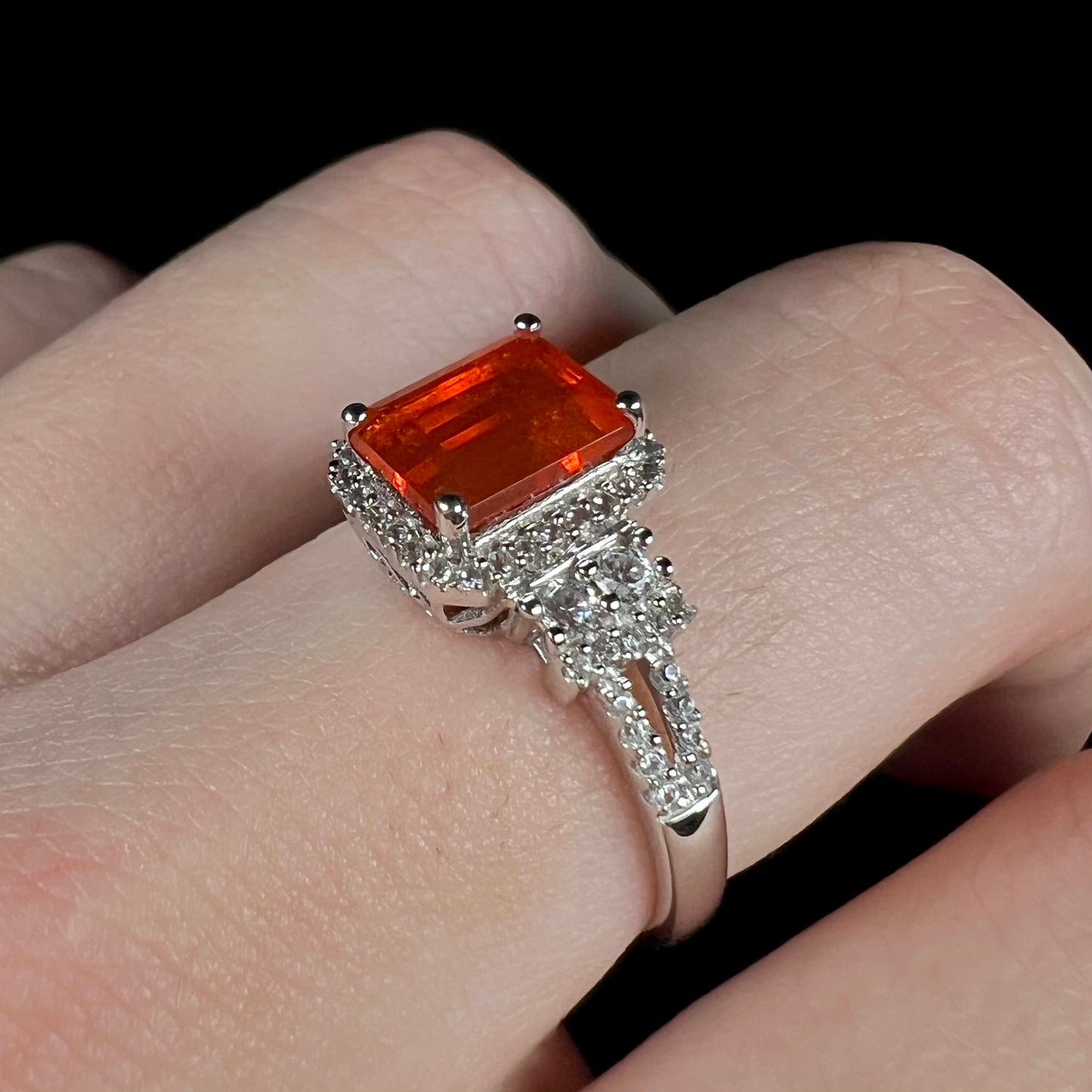Angled close-up of a fire opal and white zircon-accented ring worn on a finger against a black background under cool LED studio lighting.