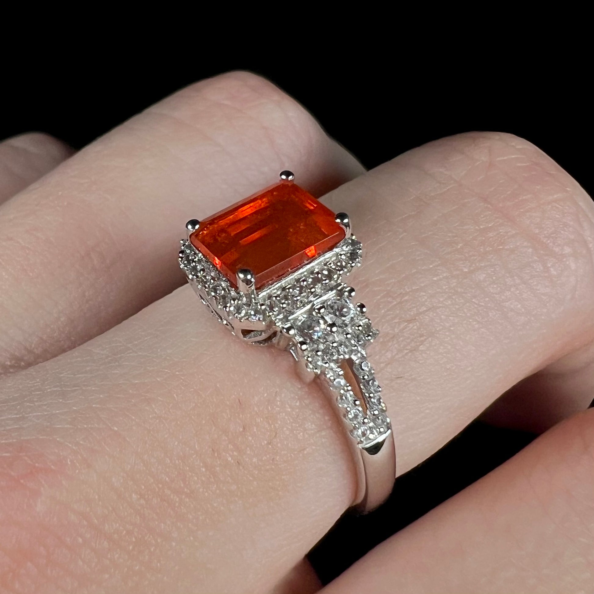 Angled close-up of a fire opal and white zircon-accented ring worn on a finger against a black background under cool LED studio lighting.