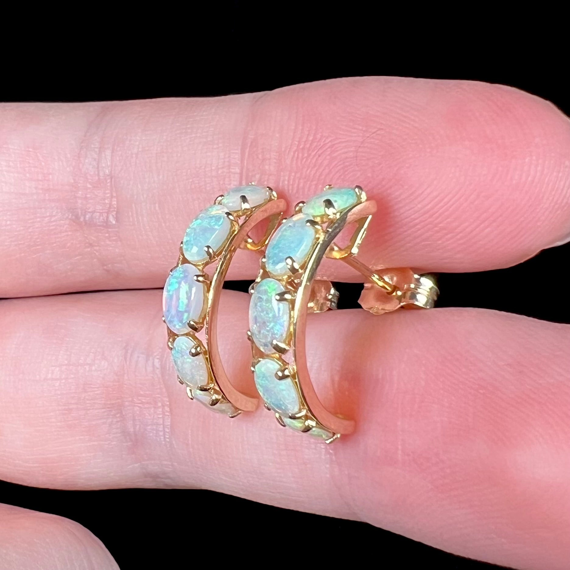 A pair of gold five-stone opal hoop earrings shown close-up from an angle held between two fingers against a black background.