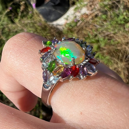 A flamboyant opal and gemstone ring shown in daylight, worn on a finger at an angle.  A rainbow of colors are seen.