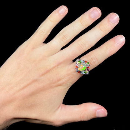 A flamboyant style opal and multi-gemstone ring, worn on a hand against a black background.