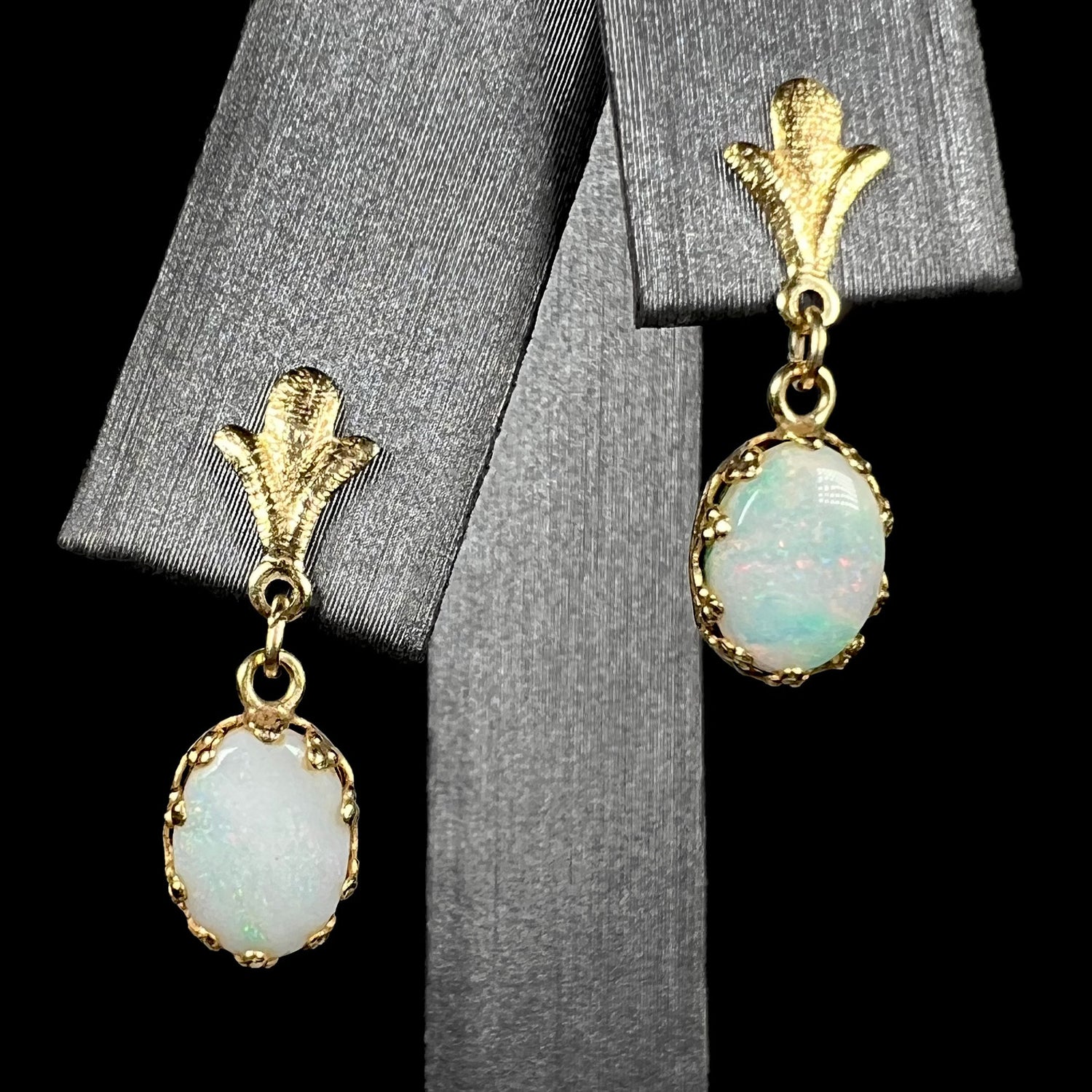 A pair of yellow gold Fleur-de-Lis Australian light opal dangle earrings shown on an earring display against a black background.