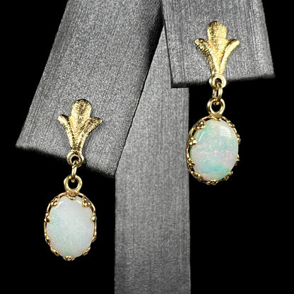 A pair of yellow gold Fleur-de-Lis Australian light opal dangle earrings shown on an earring display against a black background.