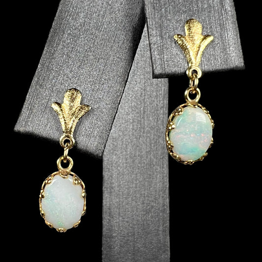 A pair of yellow gold Fleur-de-Lis Australian light opal dangle earrings shown on an earring display against a black background.