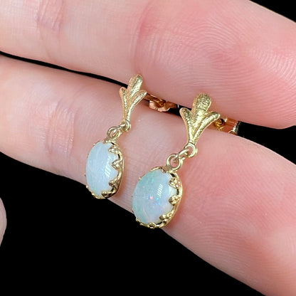 A pair of yellow gold Fleur-de-Lis Australian light opal dangle earrings shown close-up from an angle, held between two fingers.