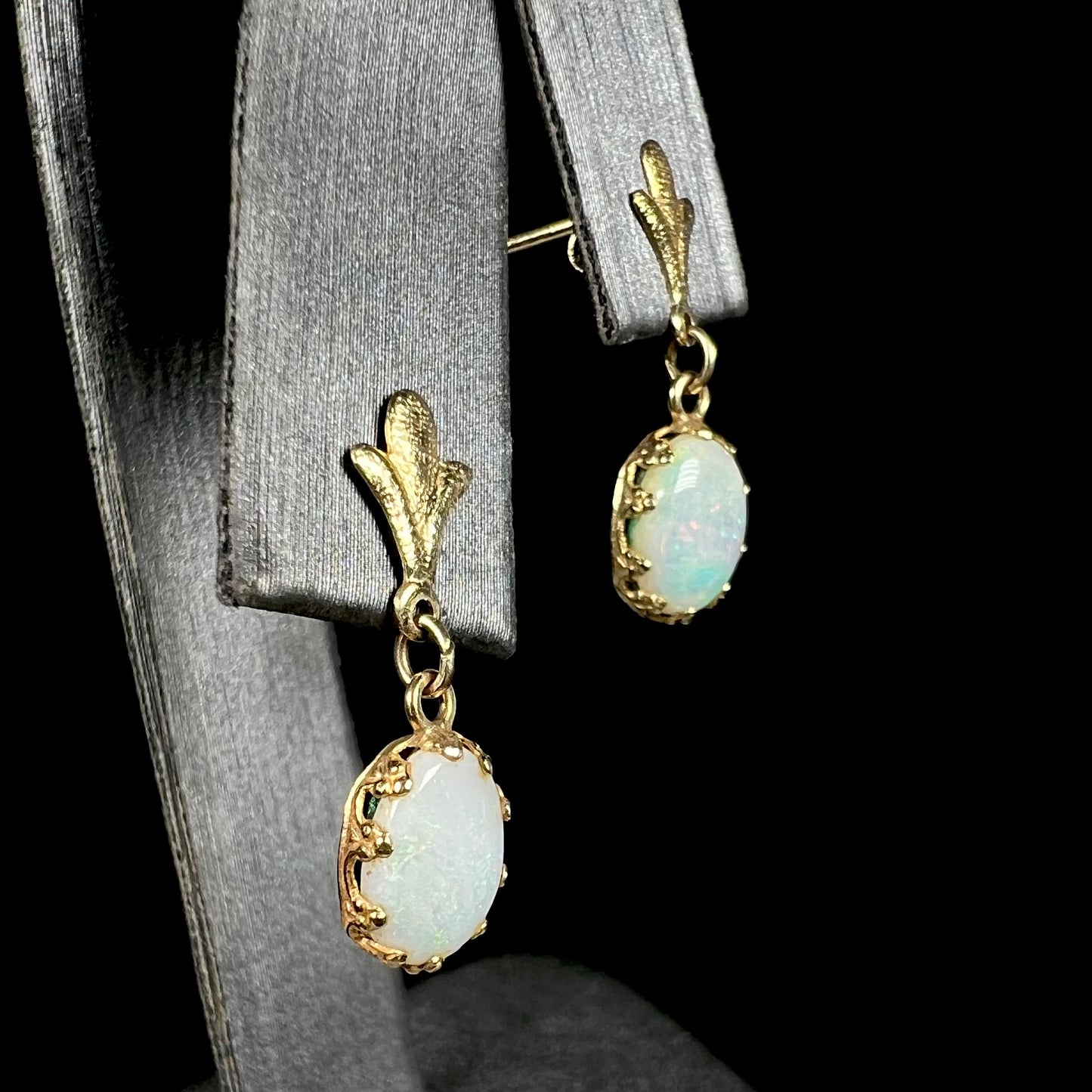 A pair of yellow gold Fleur-de-Lis Australian light opal drop earrings shown on an earring display from an angle against a black background.
