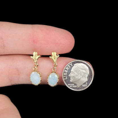 A pair of yellow gold Fleur-de-Lis Australian light opal dangle earrings shown held between two fingers next to a dime coin.