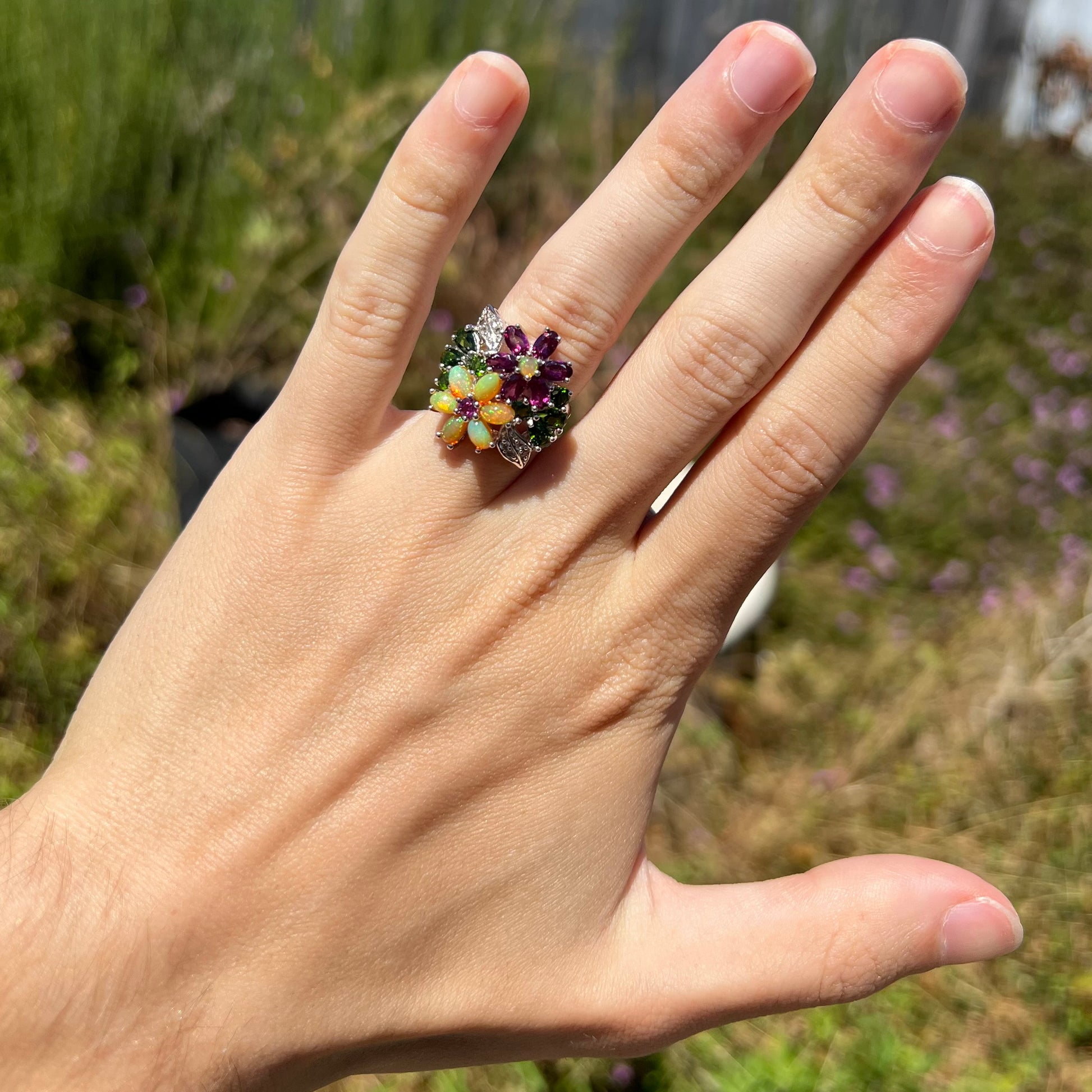 A flower ring set with different opal, garnet, and chrome diopside gemstones worn on a hand in the sunlight.