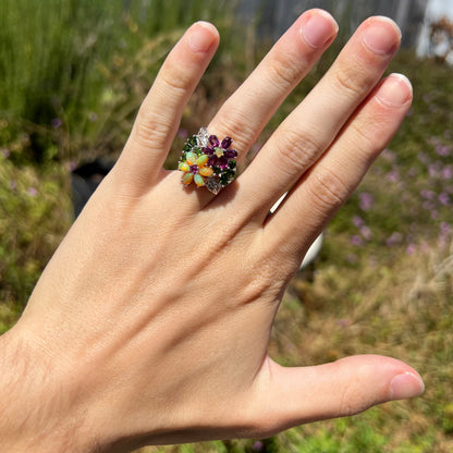 A flower ring set with different opal, garnet, and chrome diopside gemstones worn on a hand in the sunlight.