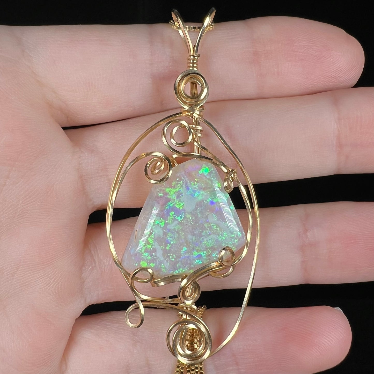 Close-up of a 13ct freeform-shaped Lambina crystal opal pendant with a gold-plated wire wrapped design, resting on fingers under cool LED light.