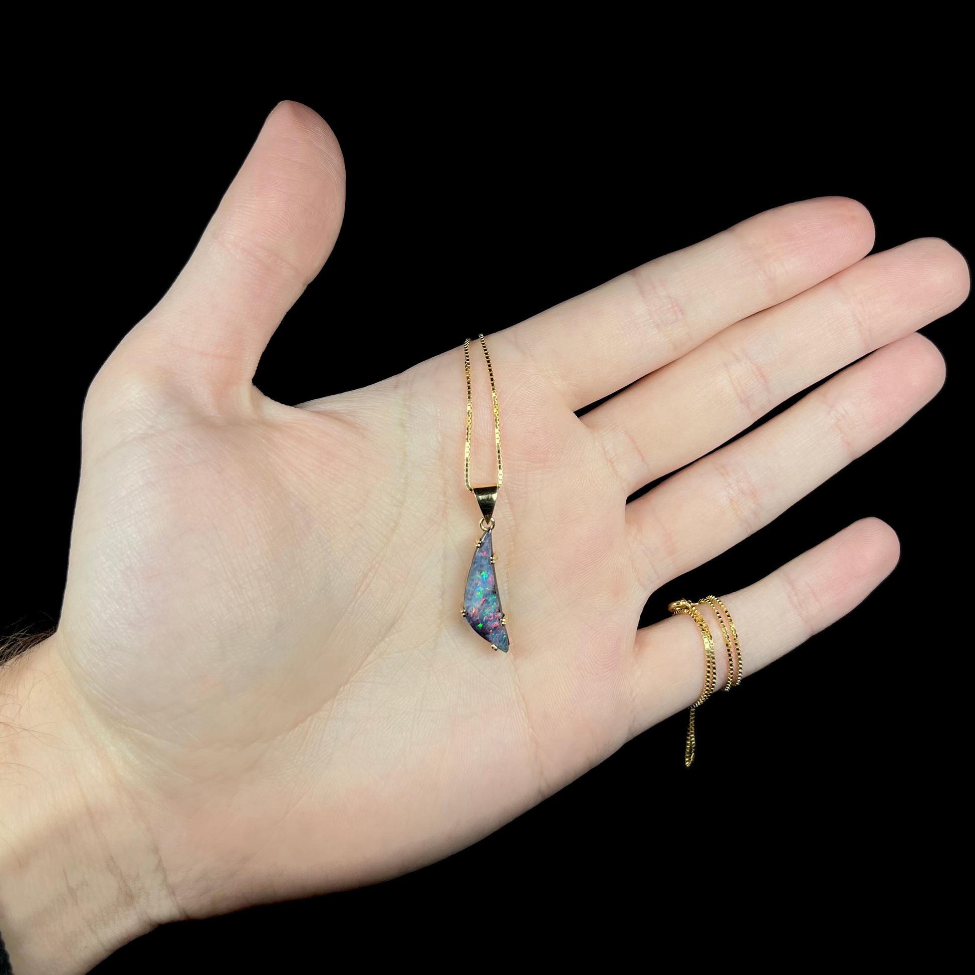 Hand holding freeform-shaped Australian boulder opal pendant in 18k gold against black background