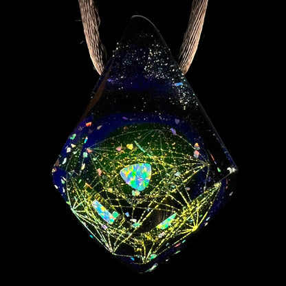 An opal in borosilicate glass pendant with a galactic-inspired motif by artist, Jes Durfee, shown close-up against a black background on a brown cord.