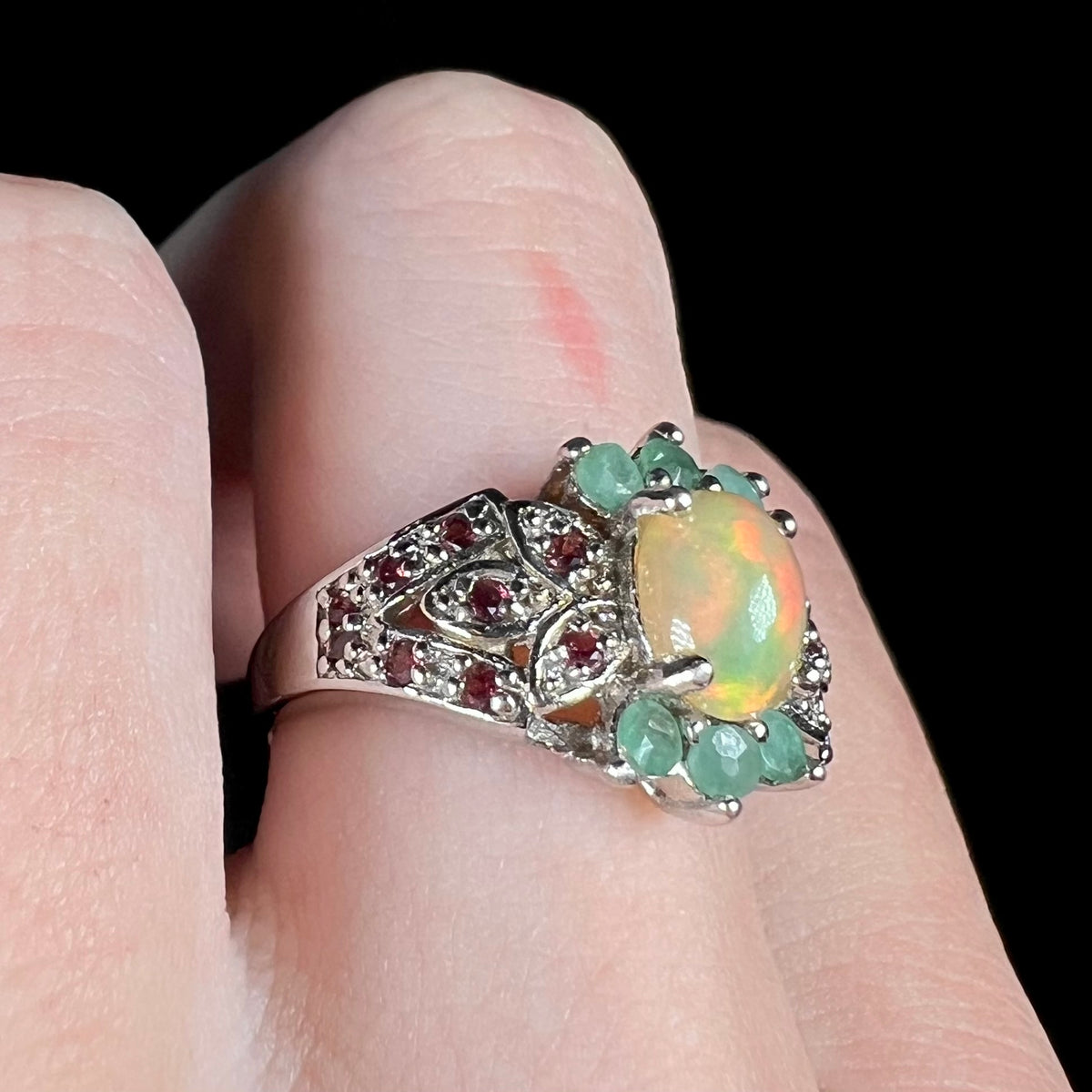 Silver garnet-accented opal ring with emerald embellishments, worn on finger