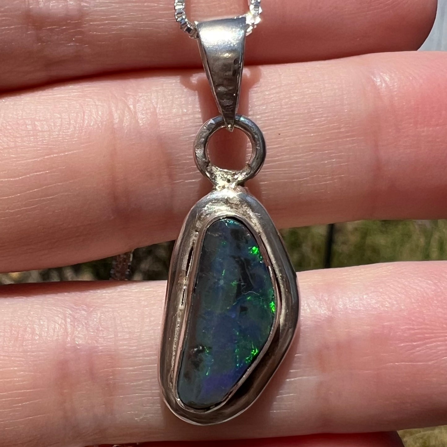 Close-up macro of a natural, genuine black opal pendant in sterling silver, held resting on fingers in sunlight.