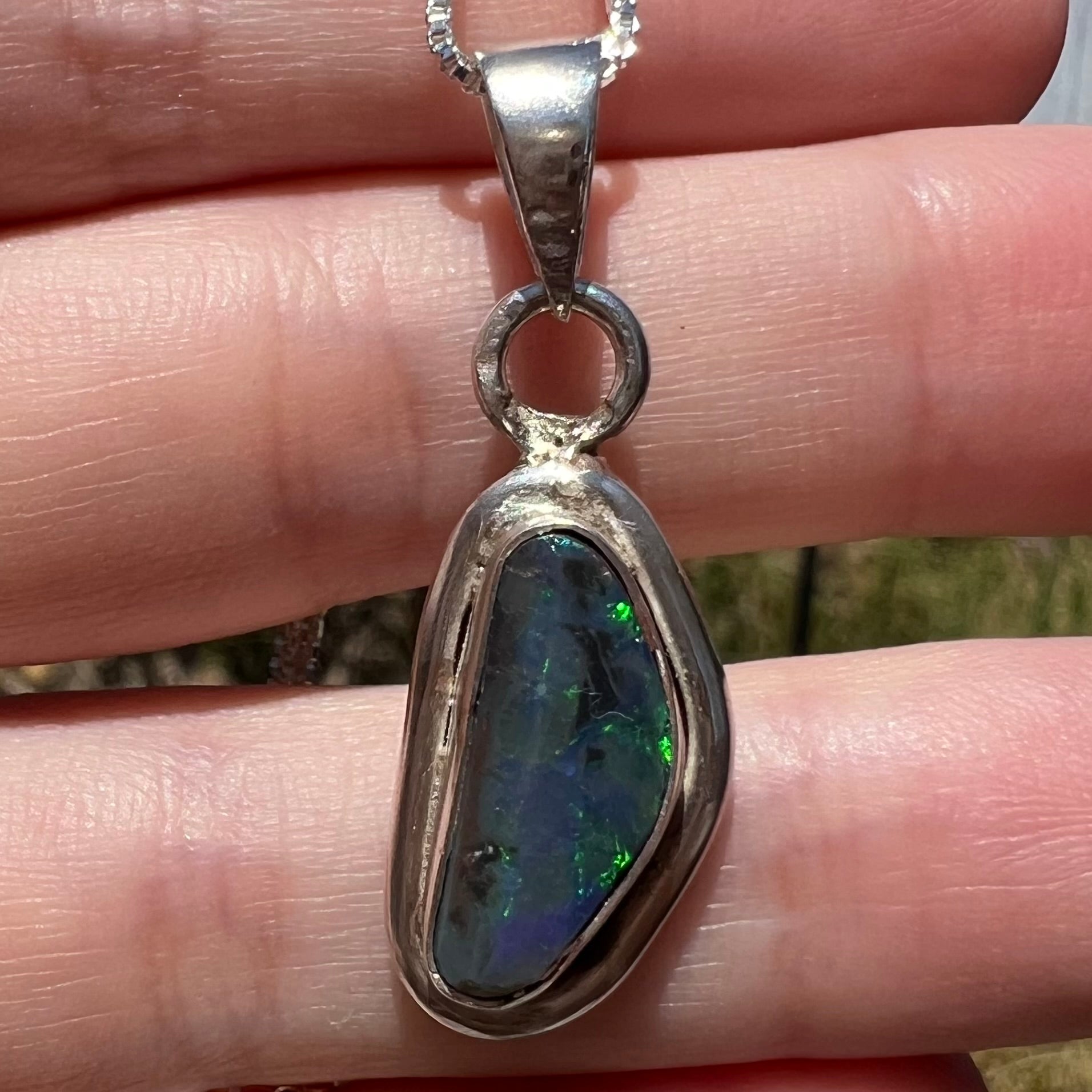 Close-up macro of a natural, genuine black opal pendant in sterling silver, held resting on fingers in sunlight.
