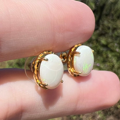 Pair of costume stud earrings set with genuine opals shown close-up in sunlight between fingers