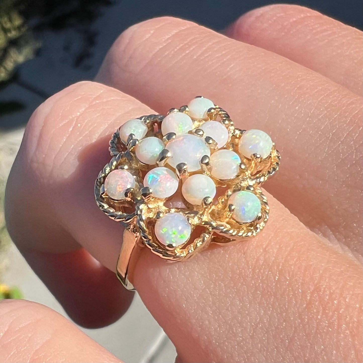 Close-up angled view of a yellow gold atomic-inspired opal cluster ring worn on a finger in the sunlight.