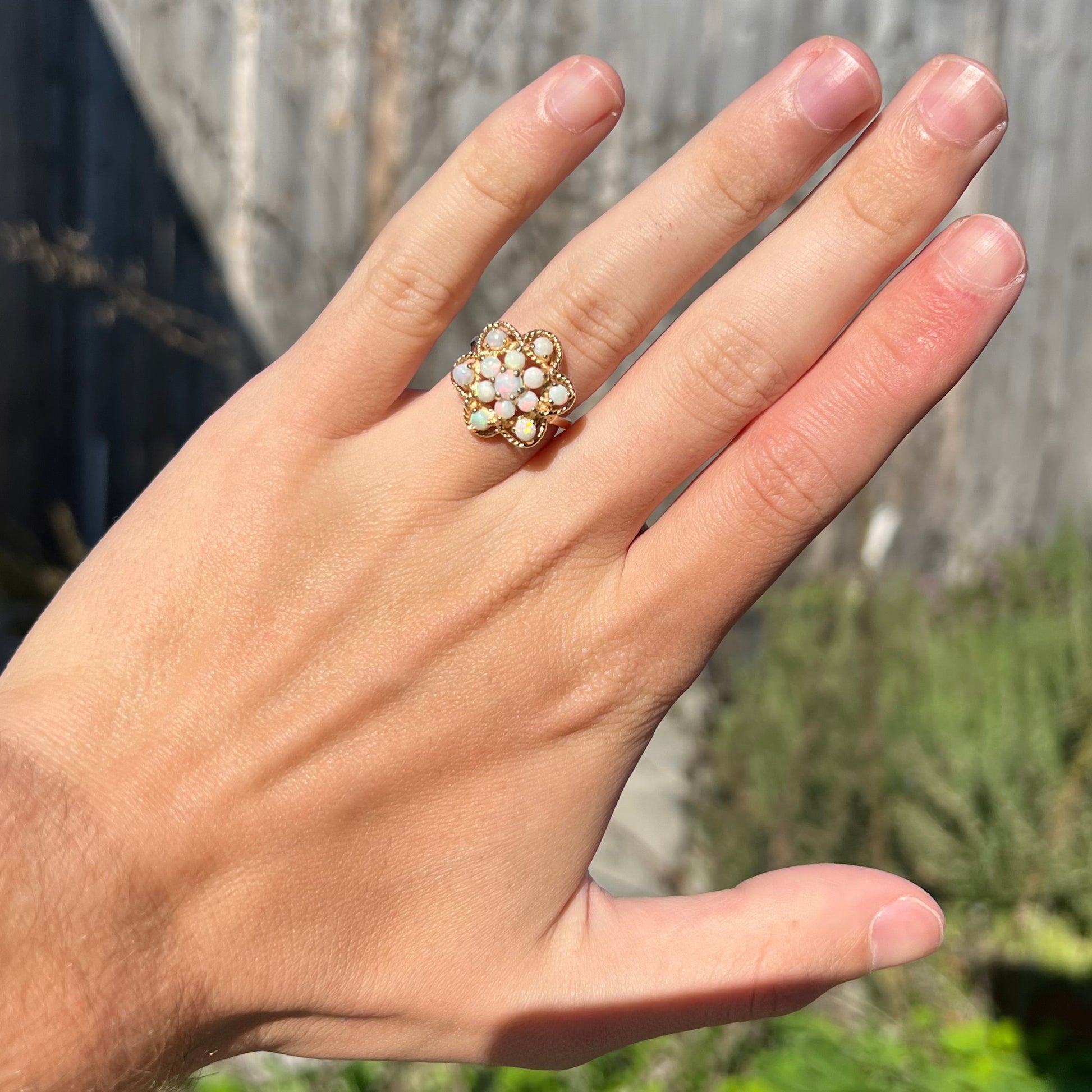 A hand wearing a yellow gold opal cluster ring with an atomic motif, shown in the sunlight.
