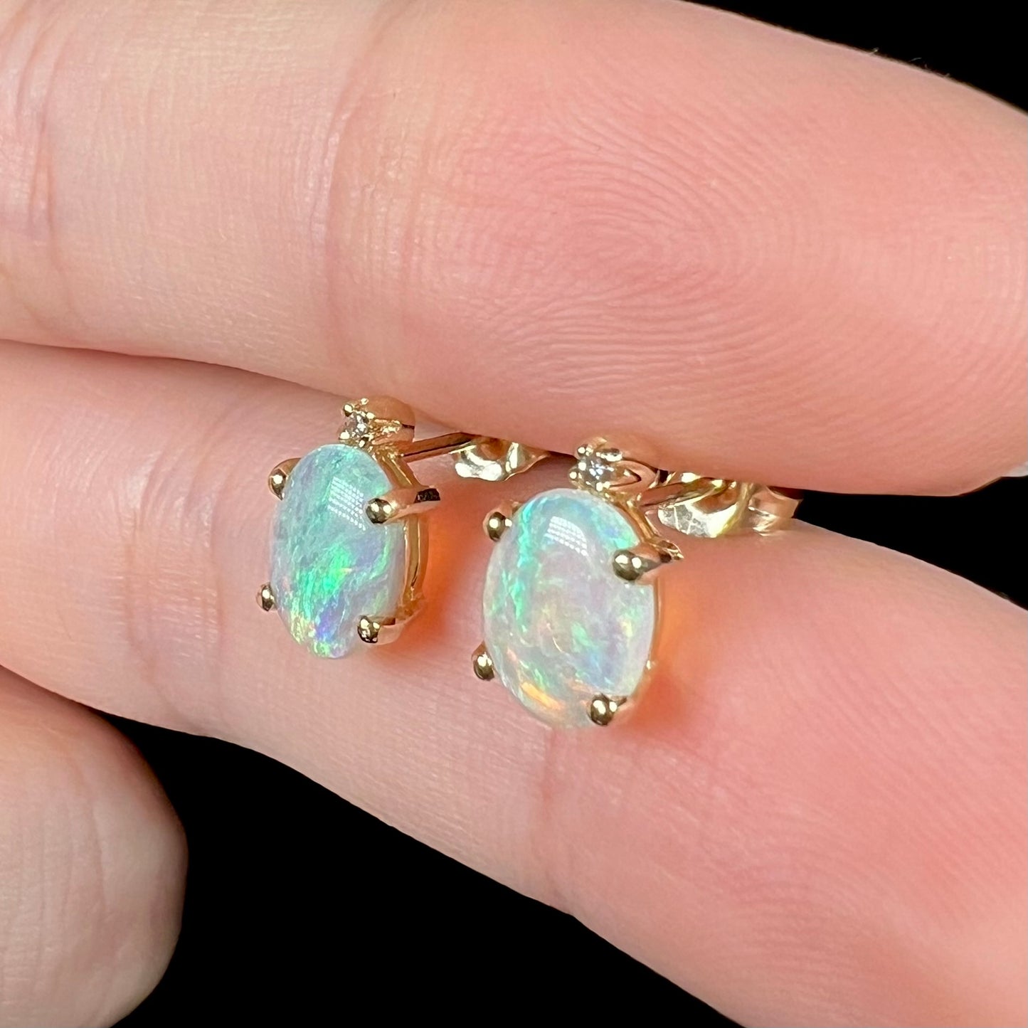 Close-up angled view of a pair of Australian crystal opal and diamond stud earrings in gold with green-blue color play, shown between two fingers.
