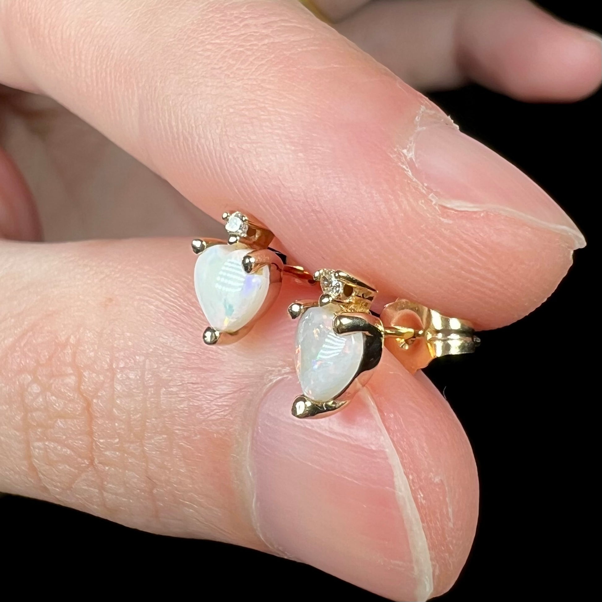 Fingers holding pair of gold heart shaped Australian opal stud earrings, showing them from side view.