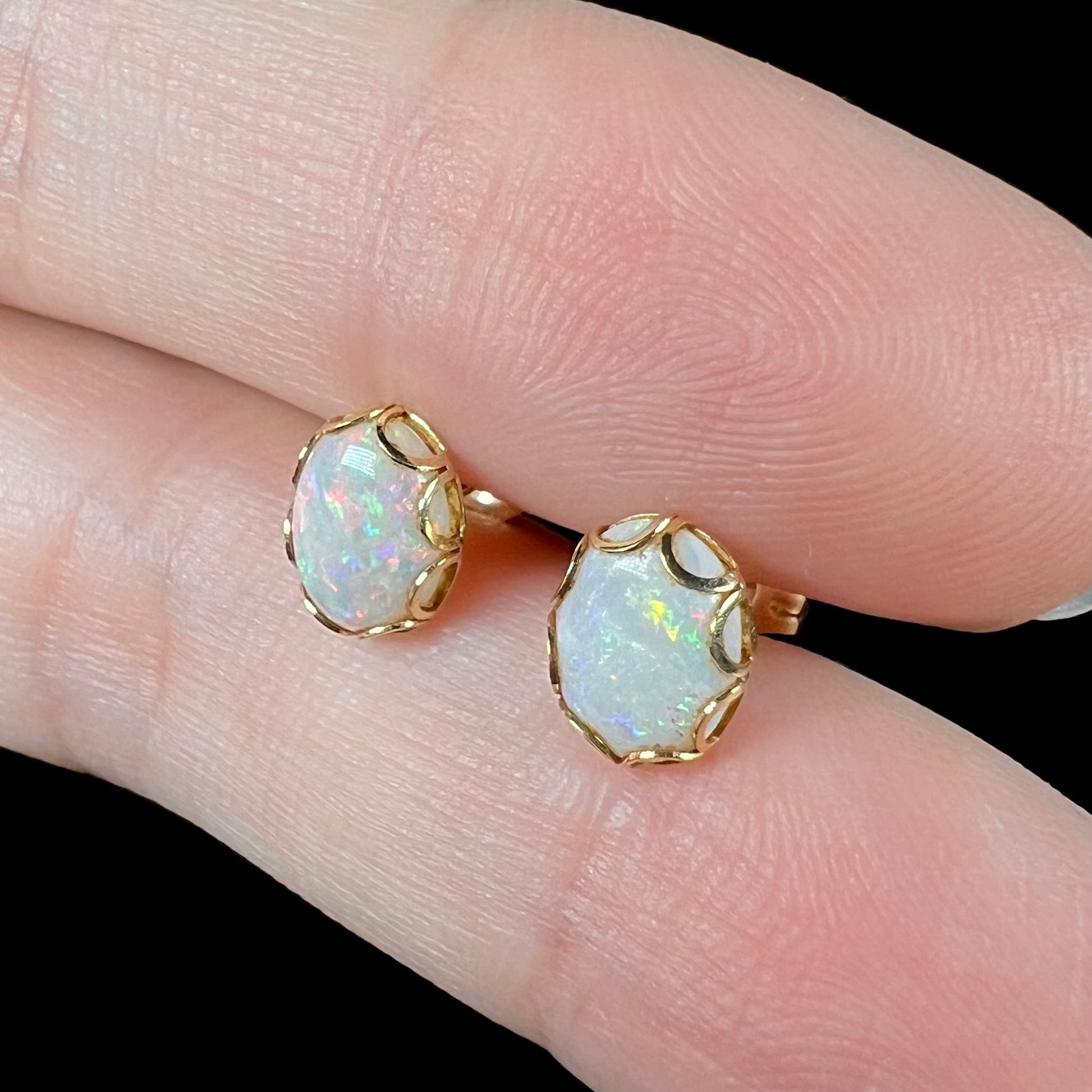 Angled close-up of a pair of Australian white opal stud earrings in yellow gold, shown between two fingers.