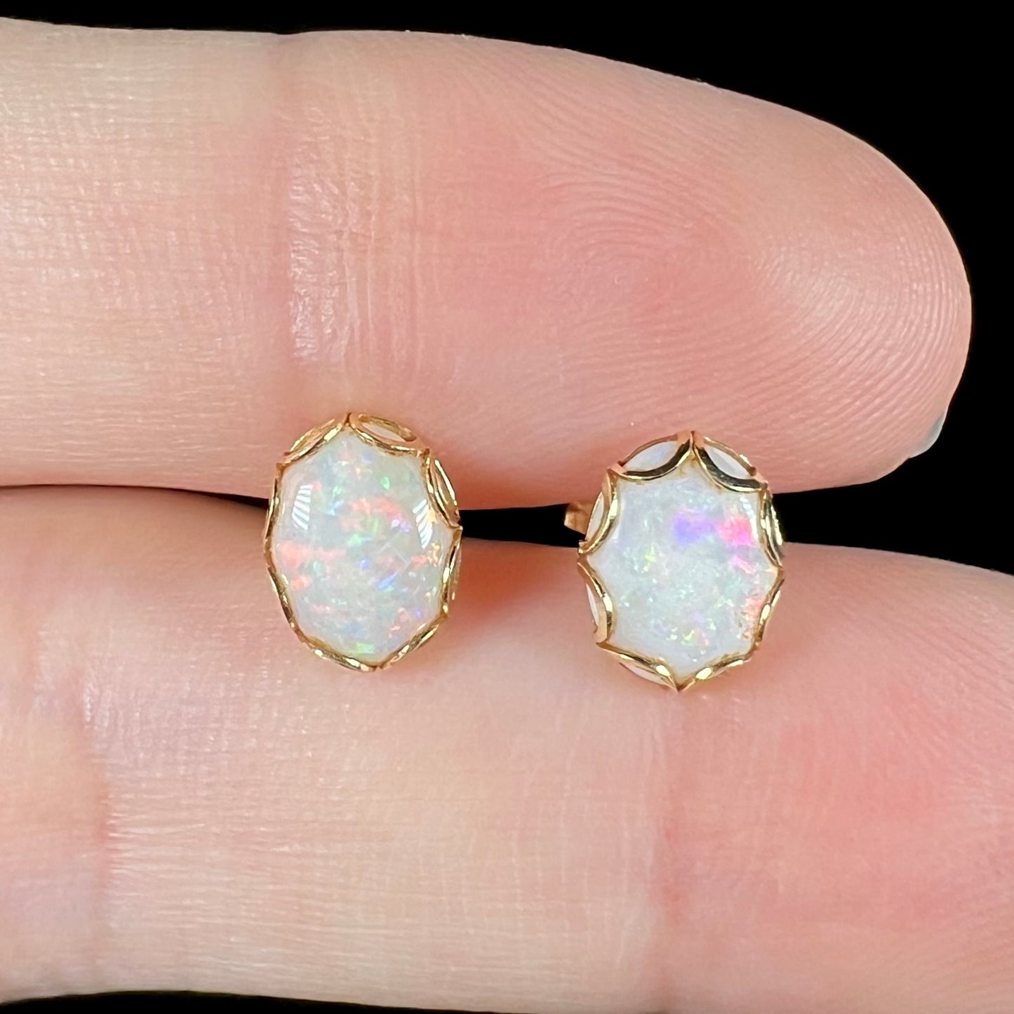 A pair of Australian white opal stud earrings in yellow gold shown close-up between two fingers.