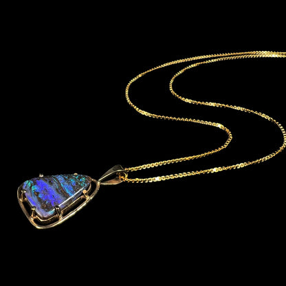 A dainty Duck Creek opalized wood necklace in 9k gold, shown from an angle laying against a black backdrop.