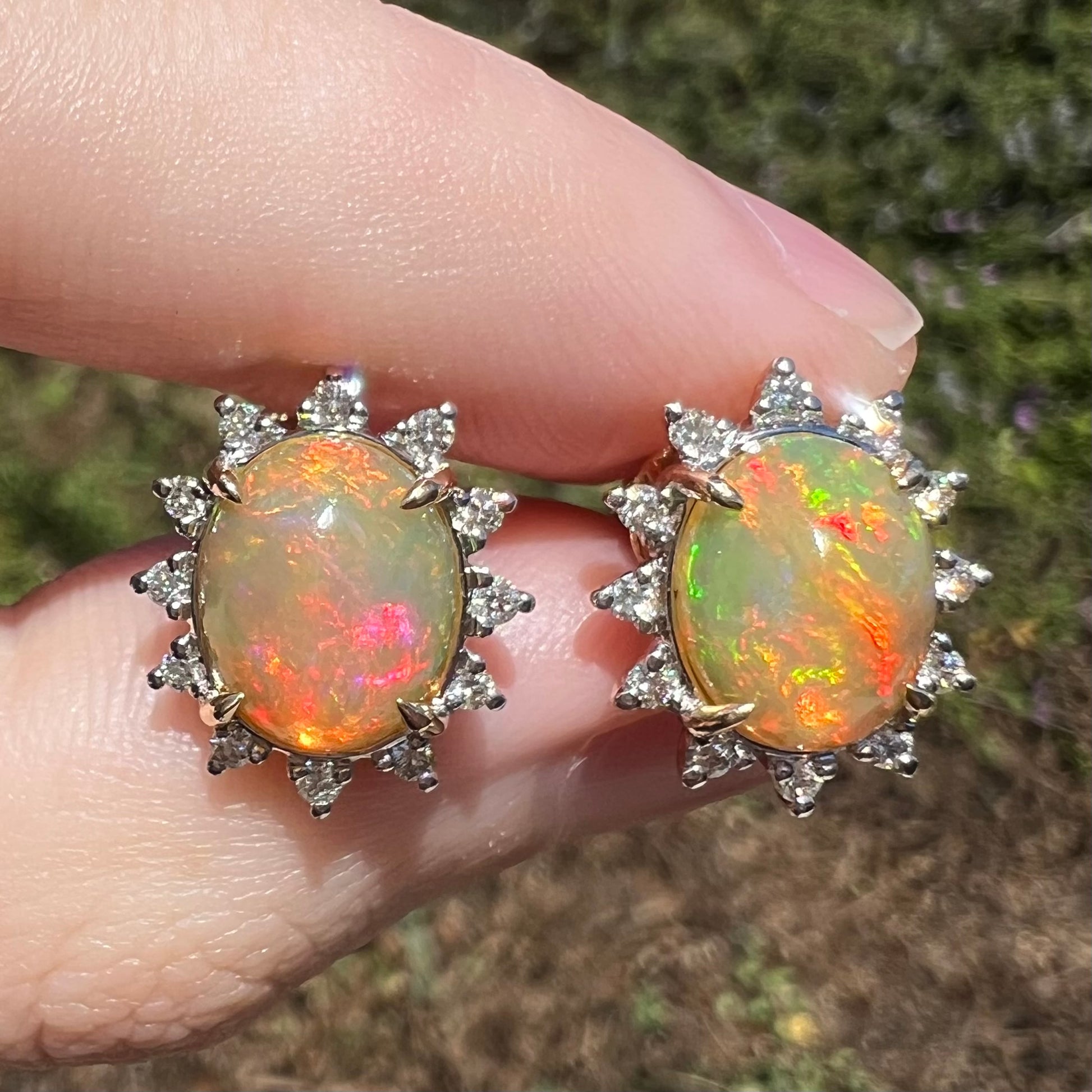 Close-up of 14k gold Ethiopian opal and diamond halo stud earrings in sunlight with red, green, and orange colors