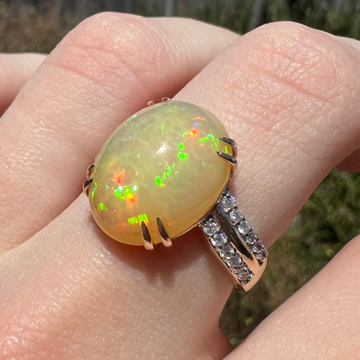 Angled close-up of women's 14k gold fire opal and diamond ring worn on finger in natural light.  Opal has green flashes of color