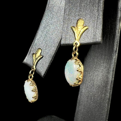 A pair of yellow gold Fleur-de-Lis Australian light opal dangle earrings shown from an angle on an earring display against a black background.