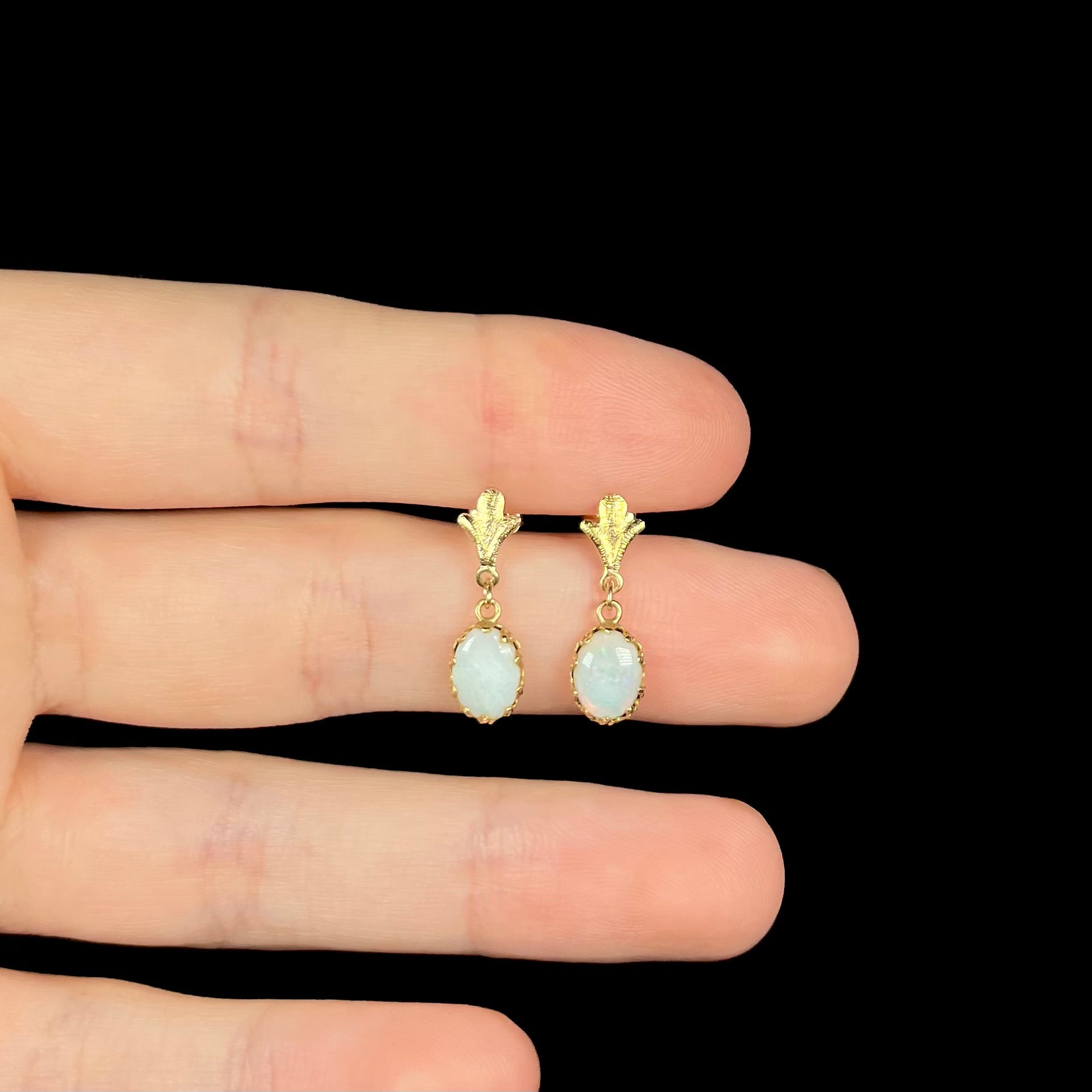 A pair of yellow gold Fleur-de-Lis Australian light opal dangle earrings held in a hand, shown against a black background.