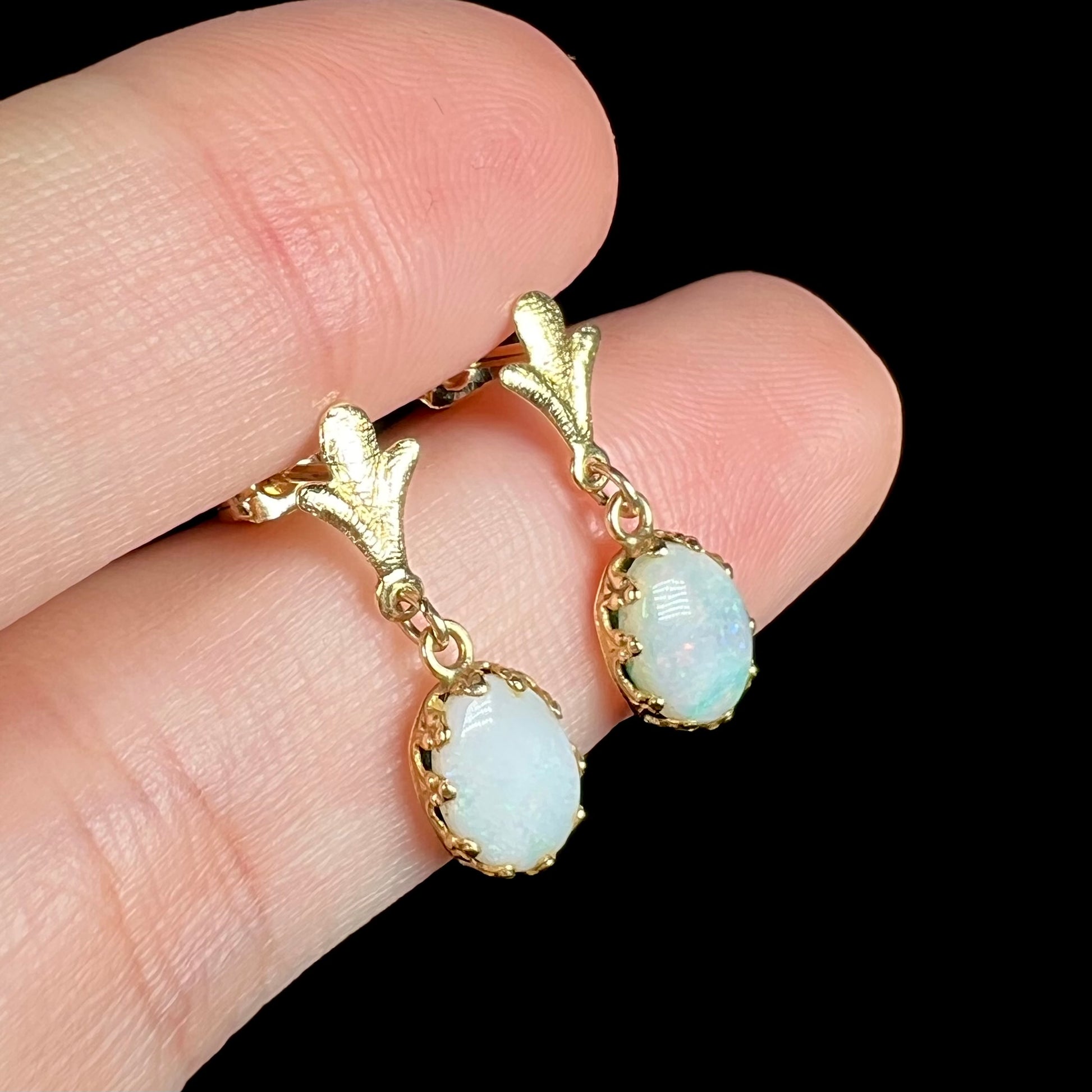 Close-up angled view of a pair of yellow gold Fleur-de-Lis Australian light opal dangle earrings, shown held between two fingers.