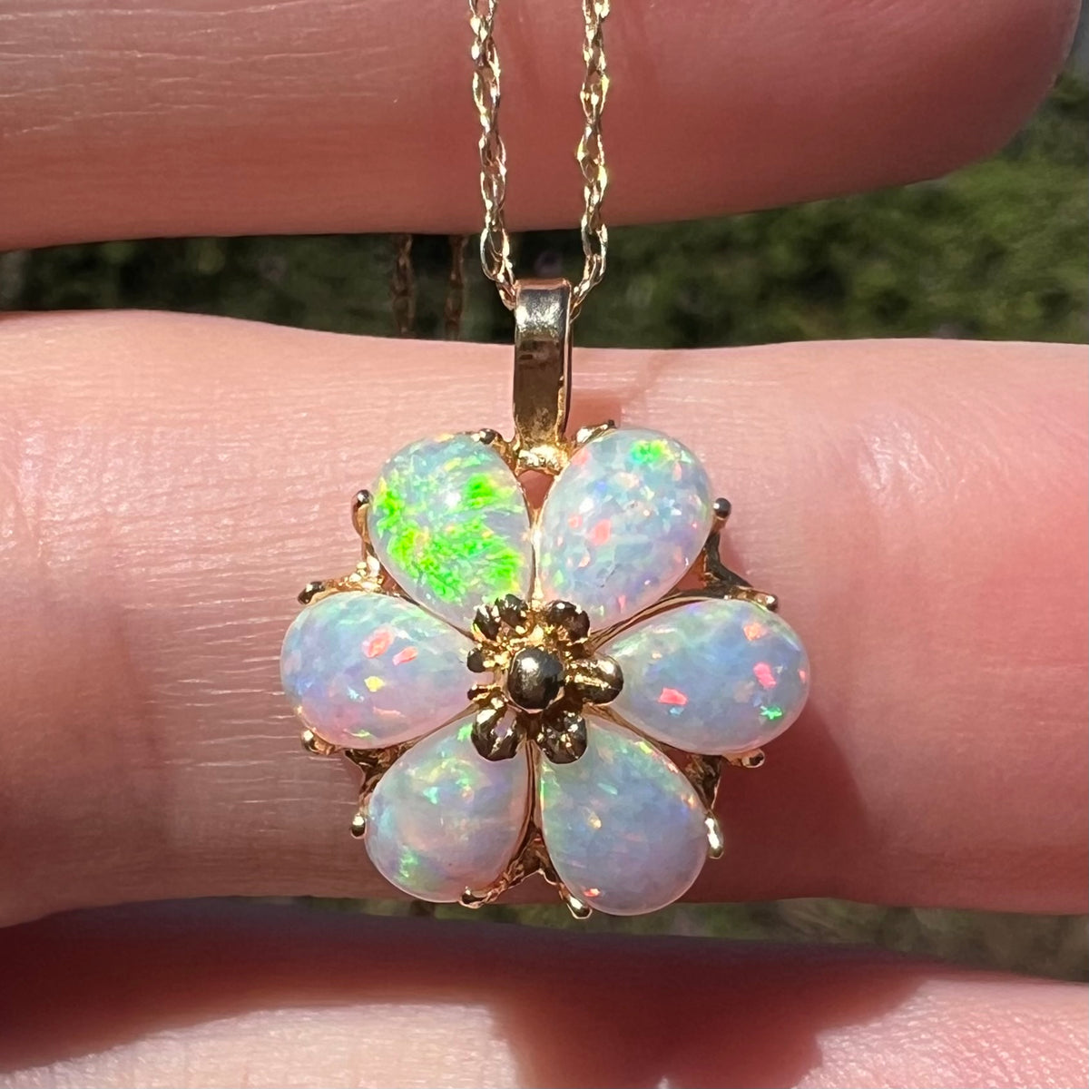 Close-up sunlit view of a 14k gold flower pendant set with six pear shaped lab-created opals, held in a hand; one opal has a bright green flash