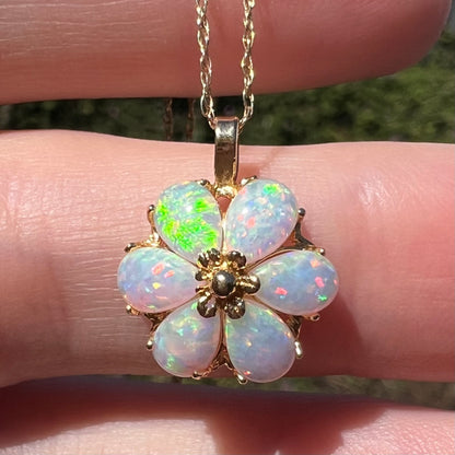 Close-up sunlit view of a 14k gold flower pendant set with six pear shaped lab-created opals, held in a hand; one opal has a bright green flash