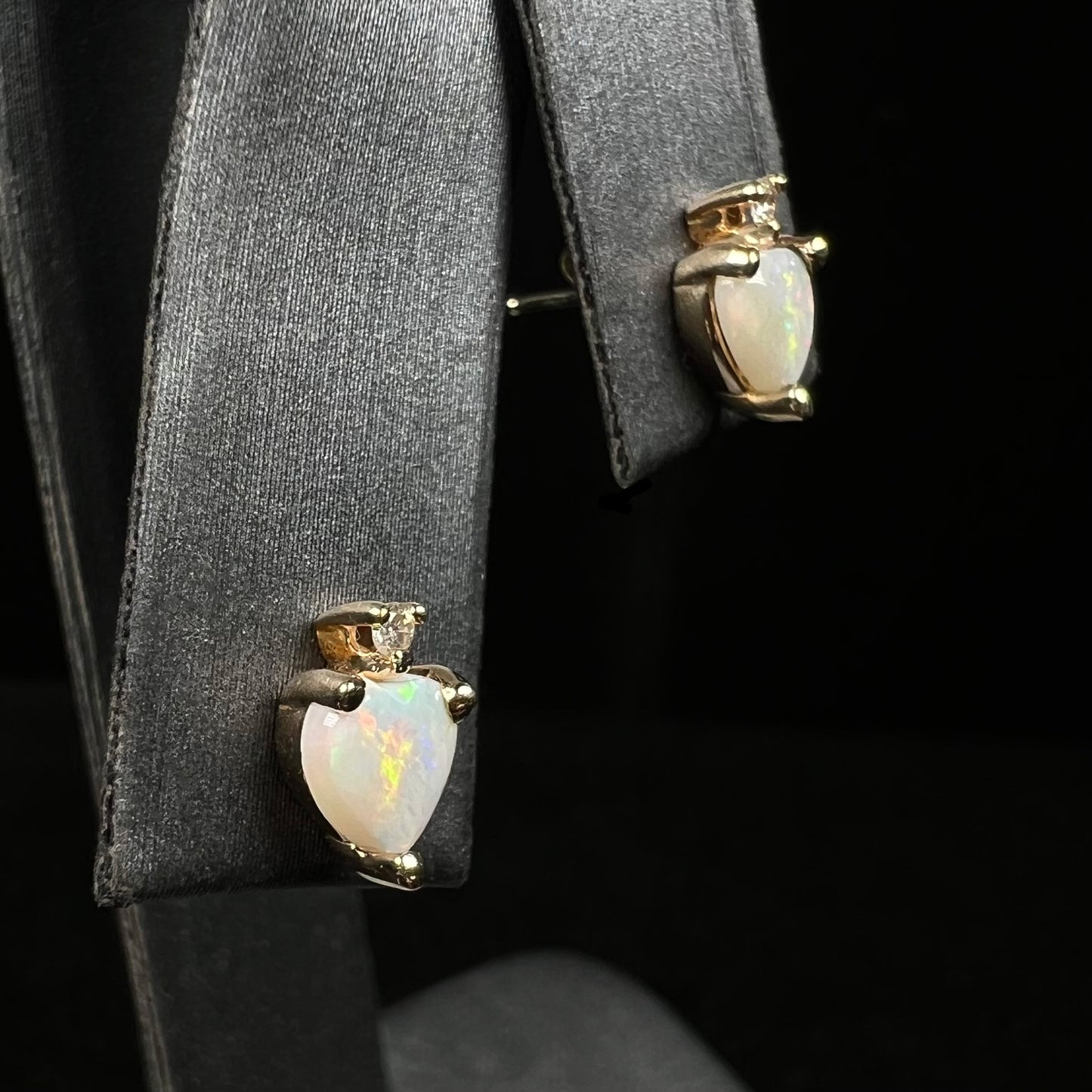 Side view of a pair of gold heart shaped opal stud earrings with diamond accents, shown on an earring display.  One opal has an orange flash of color.
