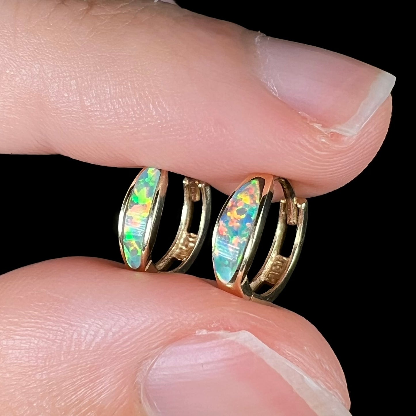 A pair of yellow gold lab-created opal inlay huggie hoop earrings shown close-up from an angle held between two fingers against a black backdrop.