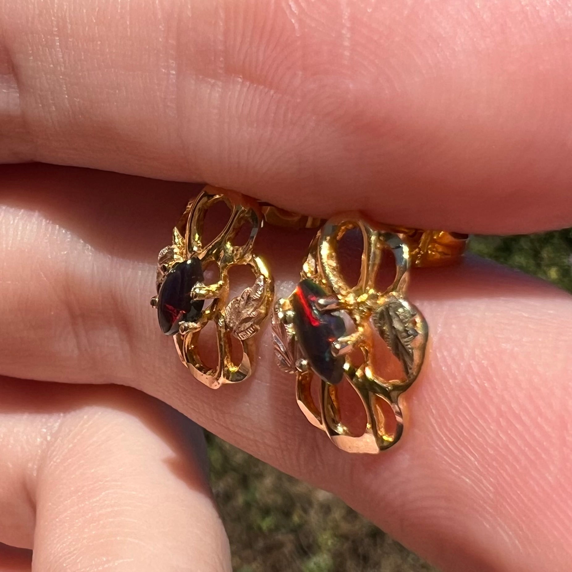 Multi-tone gold grape leaf design black opal earrings, held between fingers in natural light