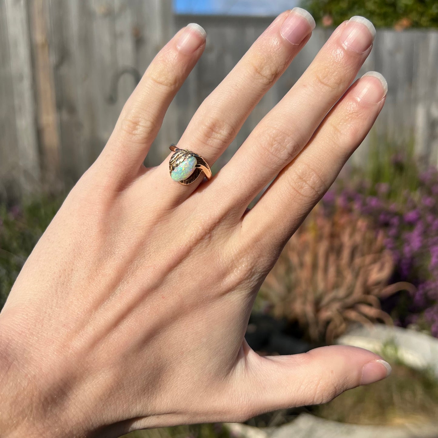 A gold ring with a leaf design and set with an opal worn on a hand outside.