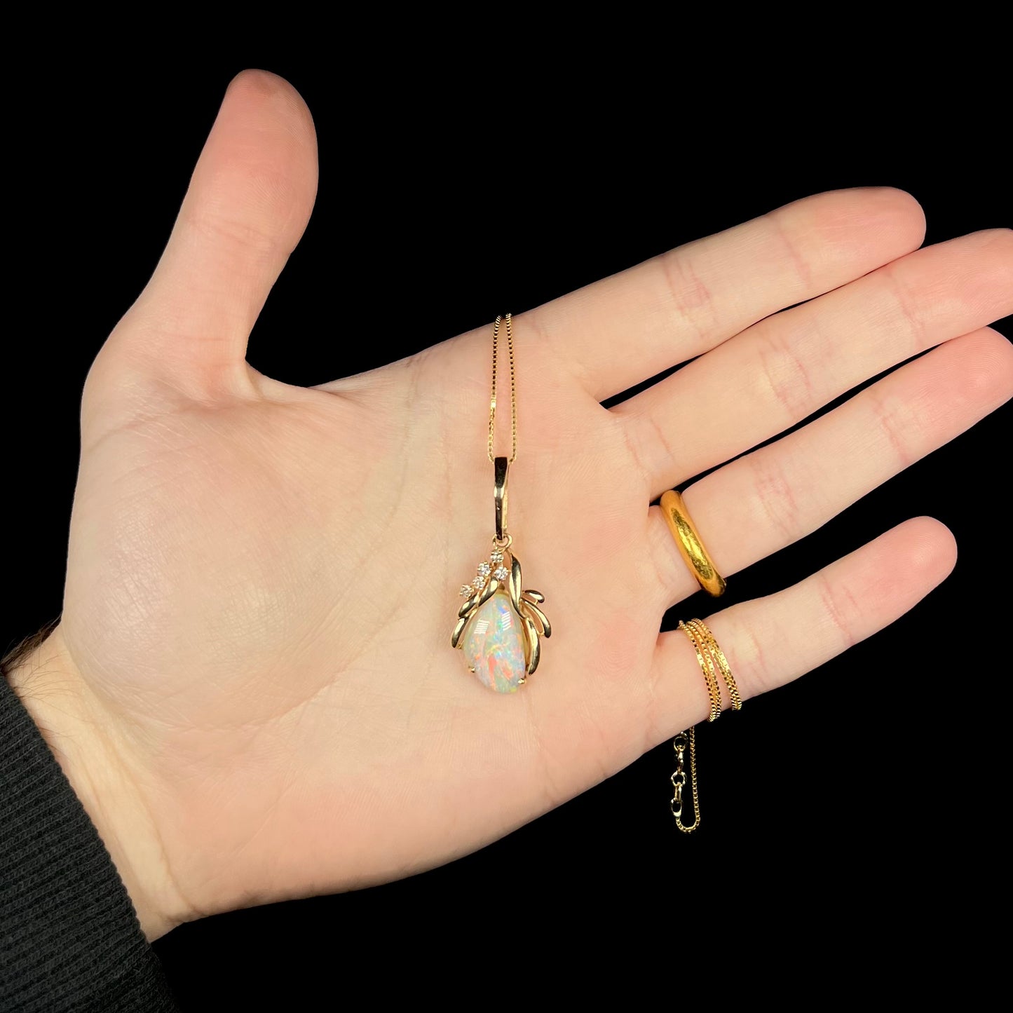 Hand holding a women's gold Modernist-style opal and diamond necklace under LED studio light.  The opal has red flashes.