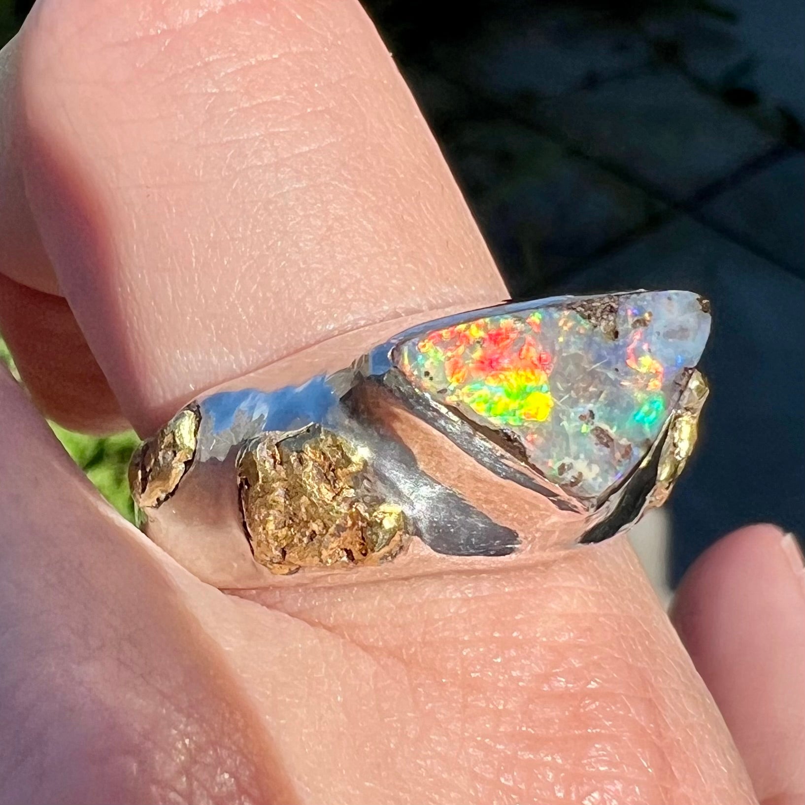 A 21k gold nugget-accented men's sterling silver boulder opal ring shown from an angle on a finger in the sunlight.  The opal has a bright red flash of color.