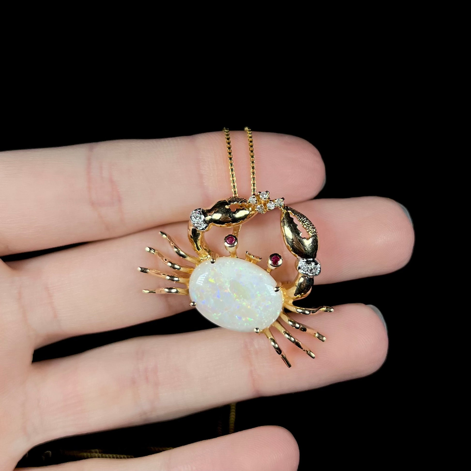 A vintage gold opal crab necklace resting on fingers against a black background, with subtle yellow opal flashes.