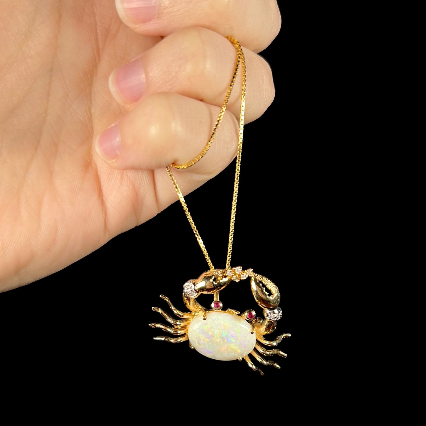 A gold opal crab necklace being dangled in a hand against a black background, displaying yellow play-of-color in the opal.